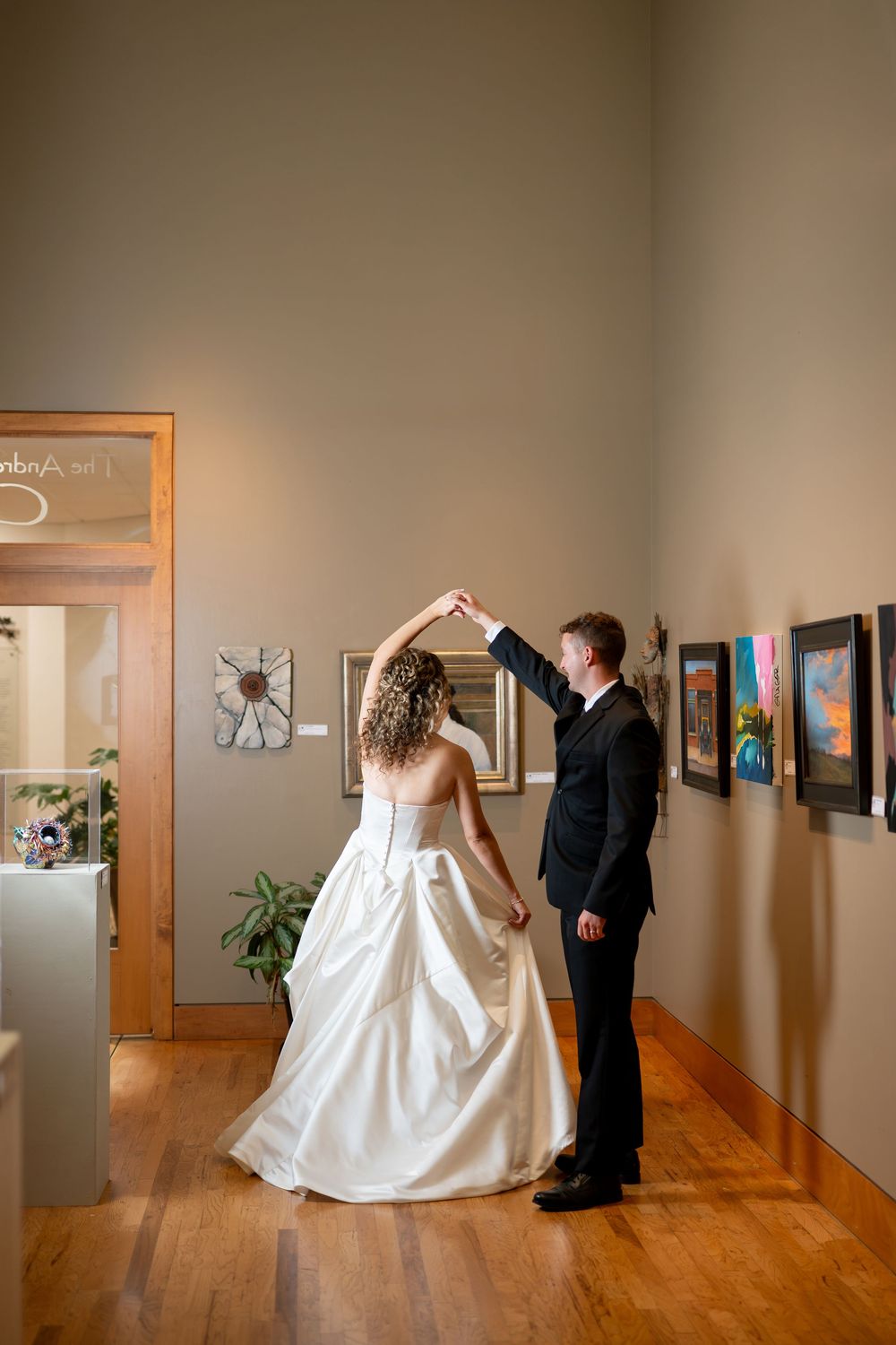 Bride and groom share a romantic dance in an art gallery, groom spinning bride in white gown on hardwood floors.