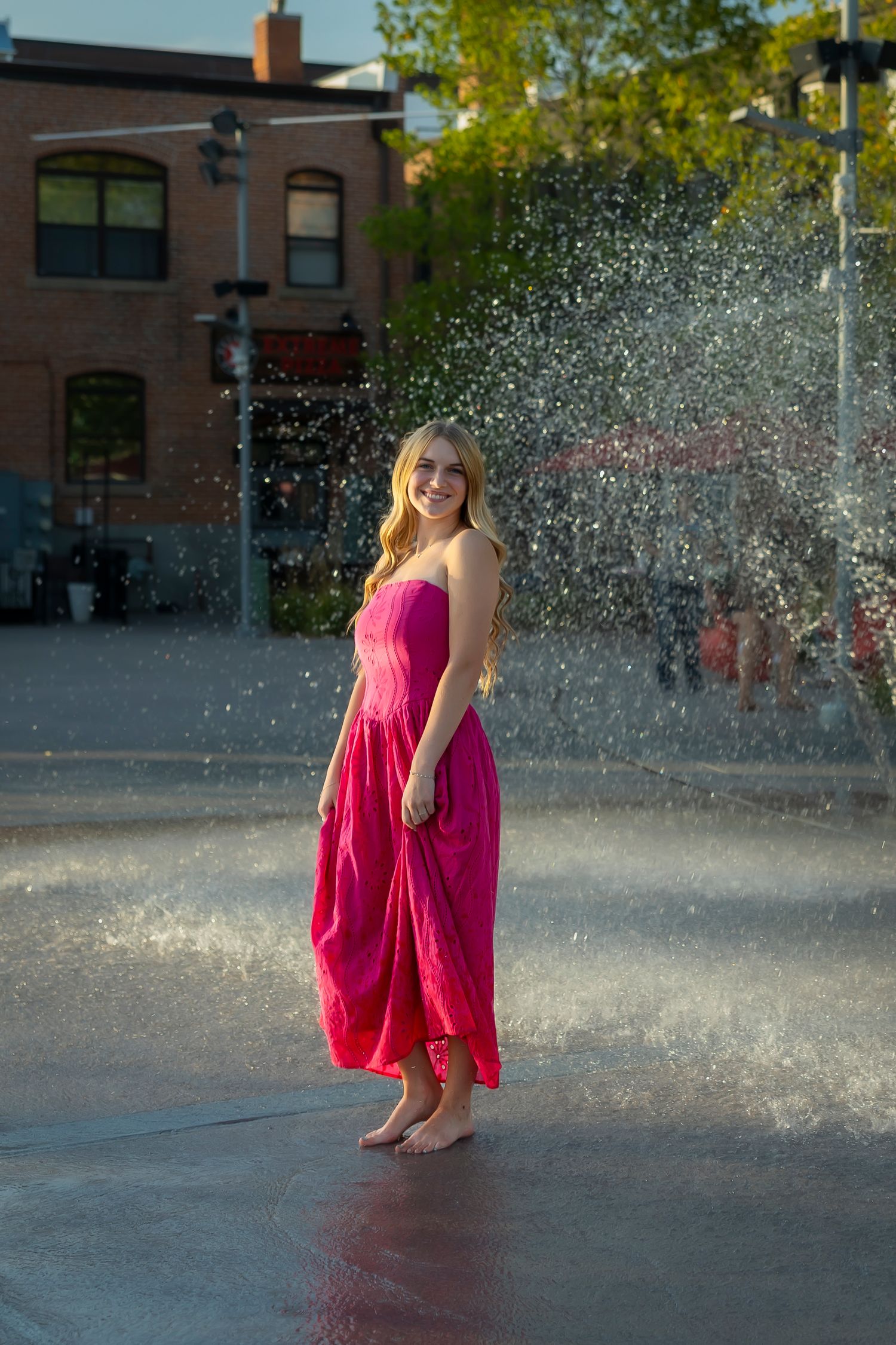 senior girl wearing a pink dress barefoot splashing around on a splash pad