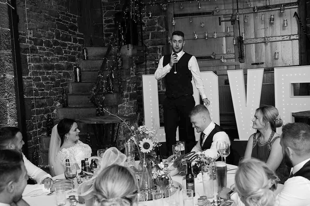 Black and white wedding reception photo showing best man giving speech beside illuminated LOVE letters at Blaithwaite House