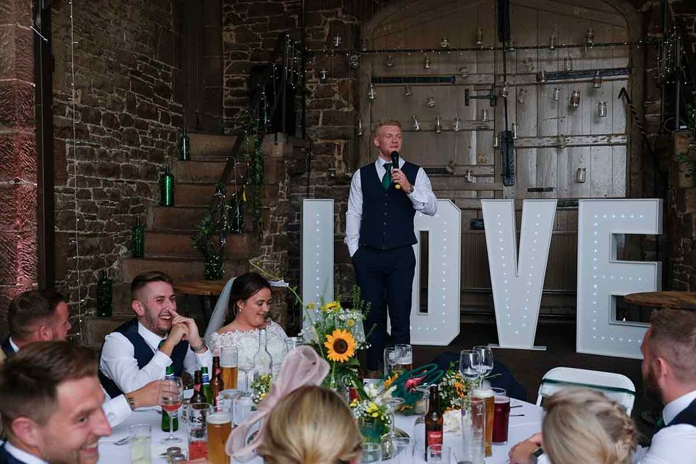 Wedding speech at Blaithwaite House with LOVE sign, sunflowers, and guests laughing at decorated reception table.