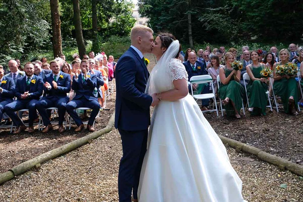 Bride and groom share first kiss at outdoor woodland wedding ceremony at romantic Blaithwaite House with seated guests in the background.
