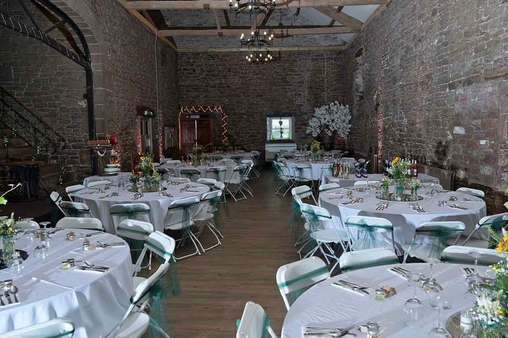 Elegant banquet hall at Blaithwaite House with brick walls, round tables draped in white linens, teal chair sashes, and chandelier lighting.