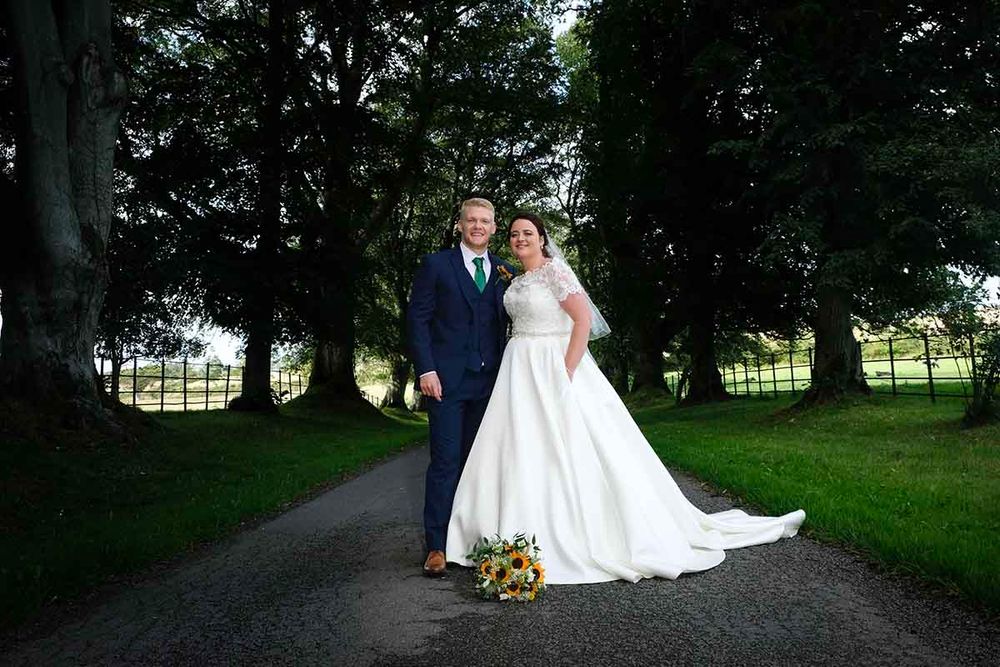 Bride in white ball gown and groom in navy suit pose on a tree-lined path at Blaithwaite House wedding venue near Wigton