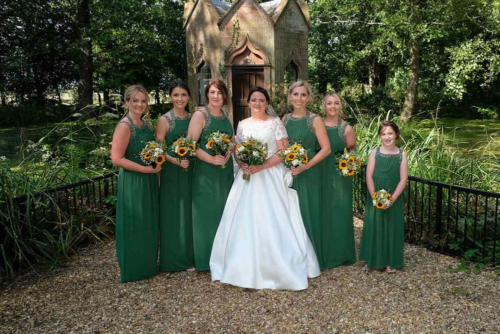 Bride in white gown with five bridesmaids in green dresses holding bouquets outside Blaithwaites famous chapel