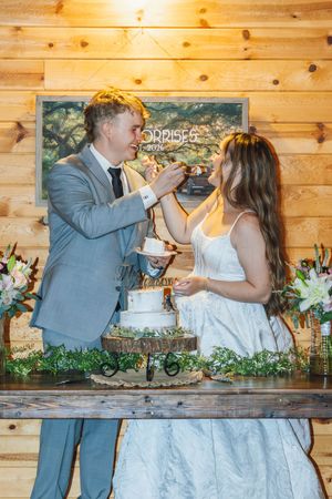 Bride and groom feeding each other wedding cake at rustic wooden venue decorated with flowers and greenery.