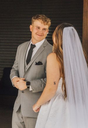 A groom in a gray suit smiles at his bride in a white wedding dress and veil during their first look ceremony.