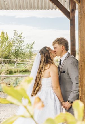 A bride and groom share a kiss under a rustic wooden pergola at an outdoor wedding venue with scenic greenery.