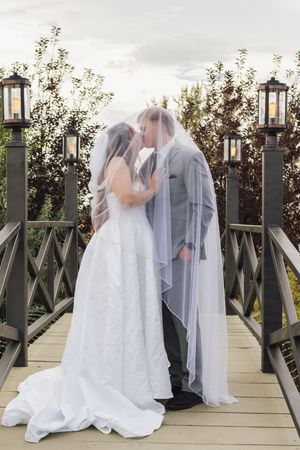 Bride and groom sharing a kiss under a flowing veil on a wooden bridge with lantern posts and lush greenery.