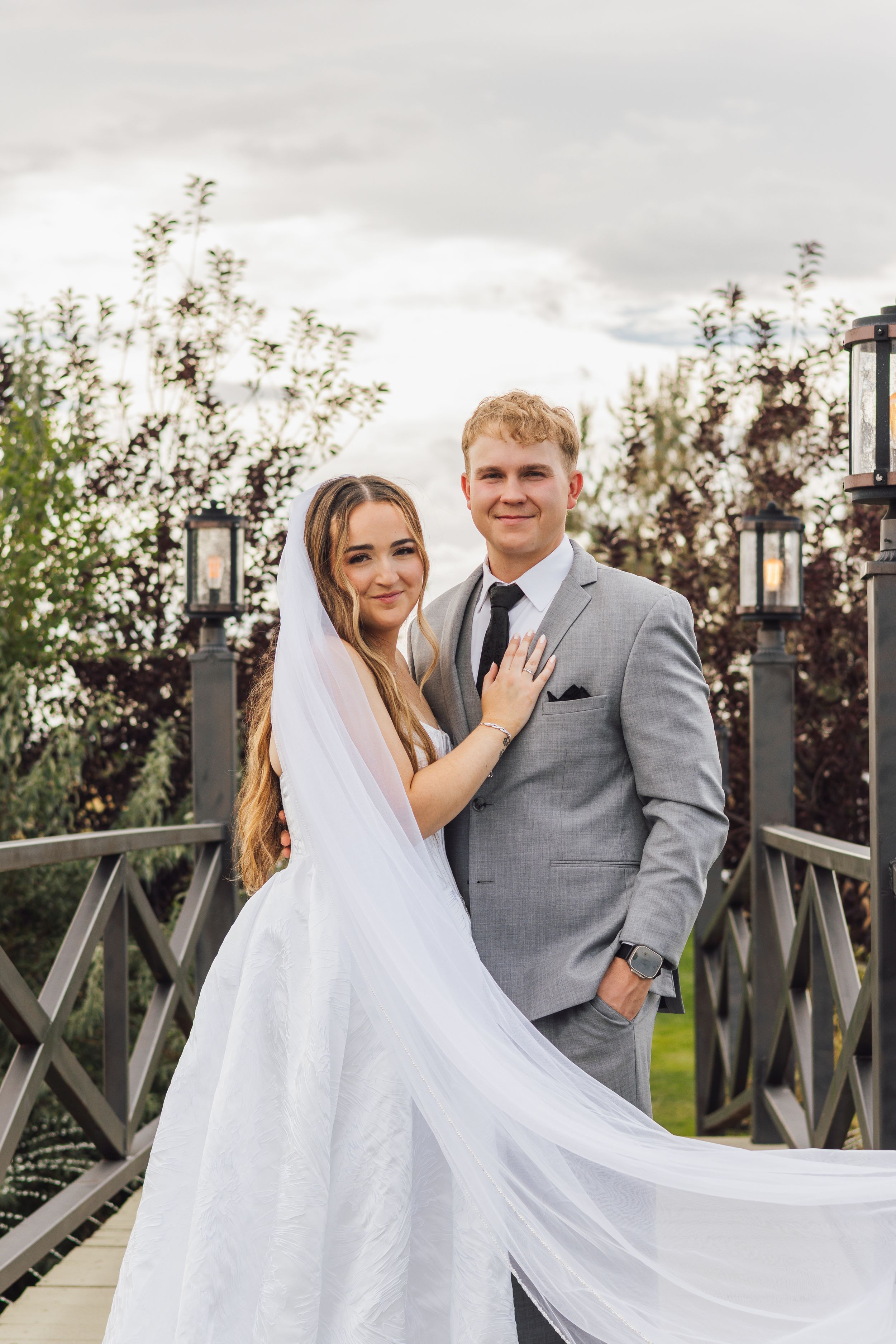 Bride and groom pose on a wooden bridge, bride showing ring, wearing white gown with veil and groom in gray suit.