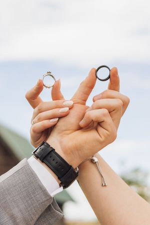 Two hands holding up wedding rings, one gold with diamonds and one black band, against a blue sky backdrop.