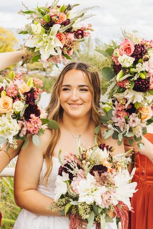 Smiling bride surrounded by colorful wedding bouquets featuring roses, dahlias, and white lilies held by bridesmaids outdoors.