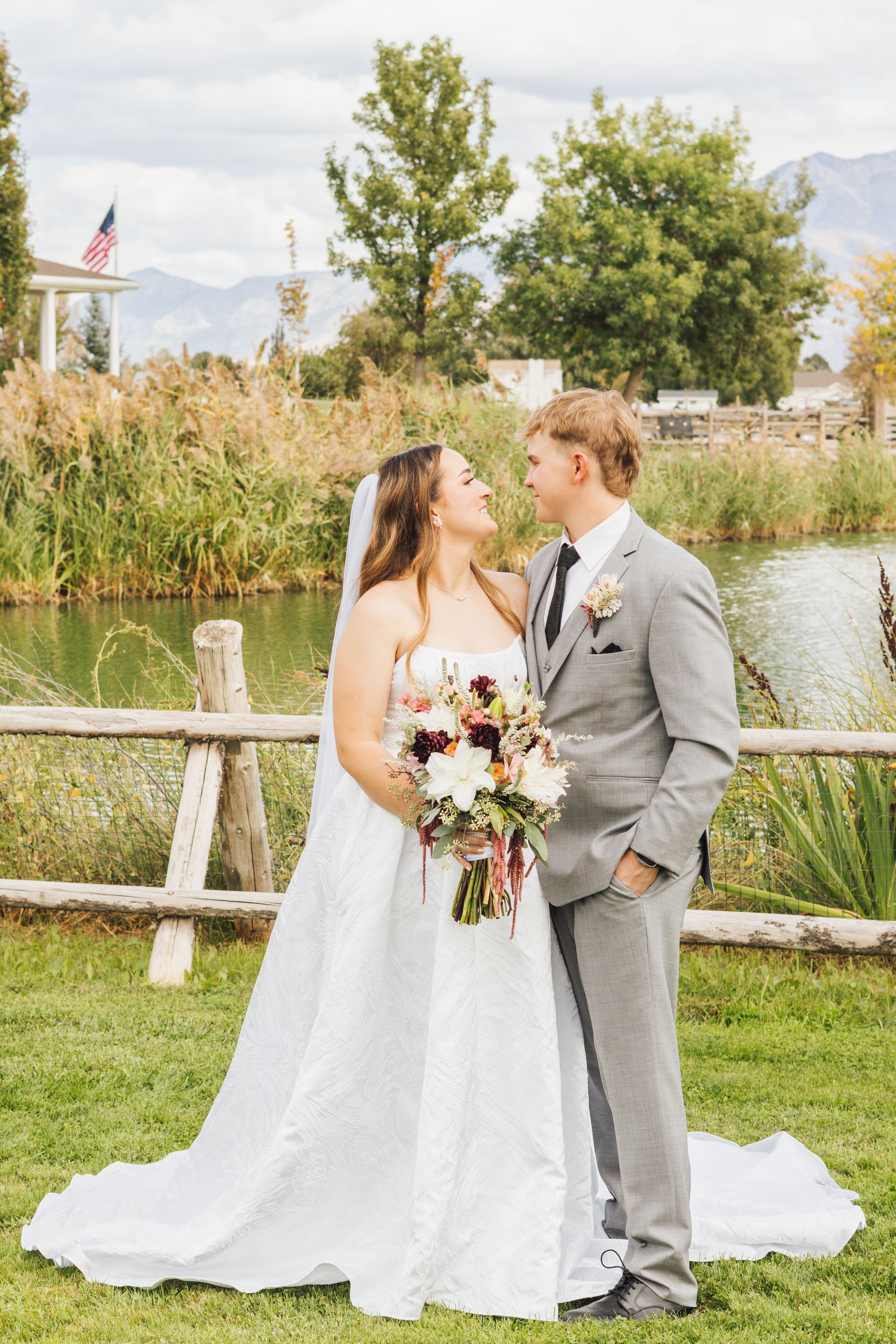 Bride and groom share a romantic moment outdoors by a pond, bride holding floral bouquet, groom in gray suit.