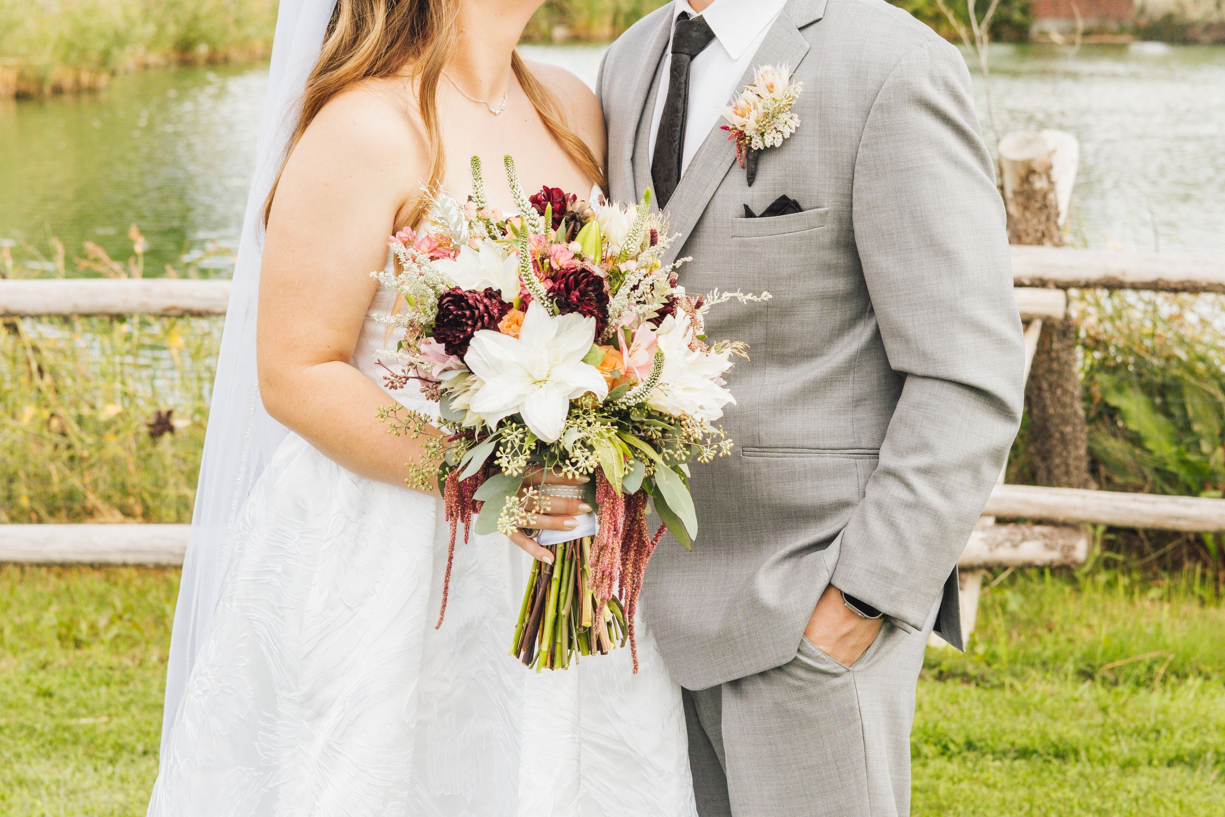 Bride and groom pose outdoors holding a lush burgundy and white wedding bouquet with greenery near a wooden fence.