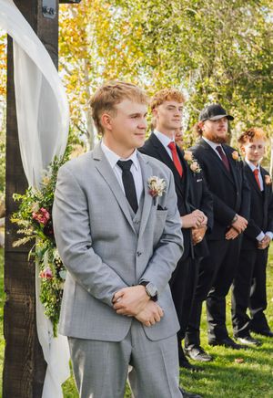 Groom in gray suit with boutonniere stands at outdoor wedding altar, groomsmen in black suits with red ties behind him.