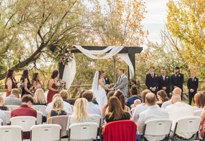 Outdoor wedding ceremony with guests seated, bridal party at floral arch, surrounded by autumn trees.