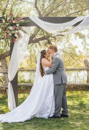 Bride and groom share first kiss under floral wedding arch with white draping and pink flowers by a serene lakeside.