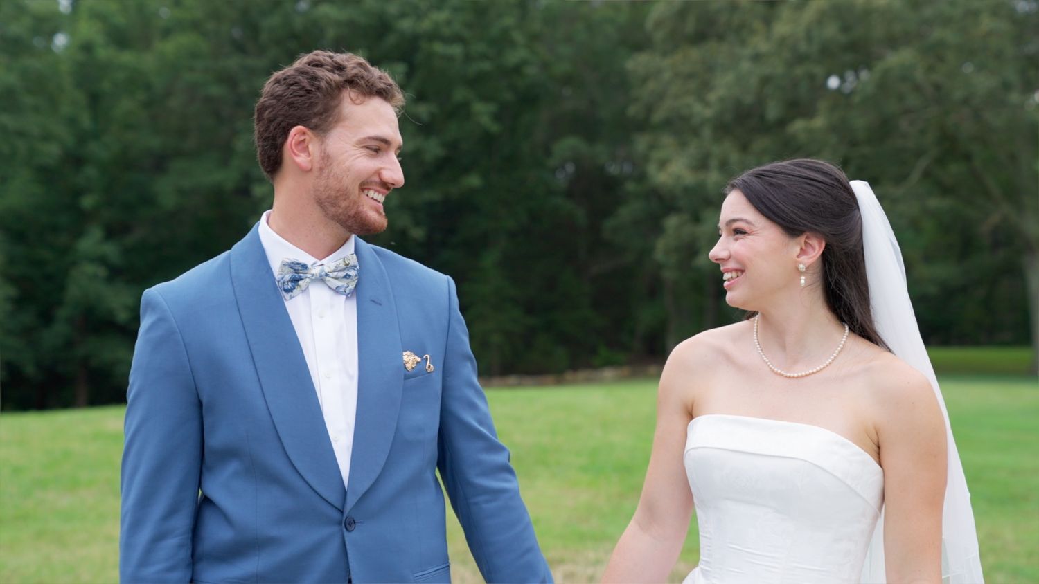 A smiling bride and groom in a blue suit and white strapless gown pose outdoors on a green lawn.