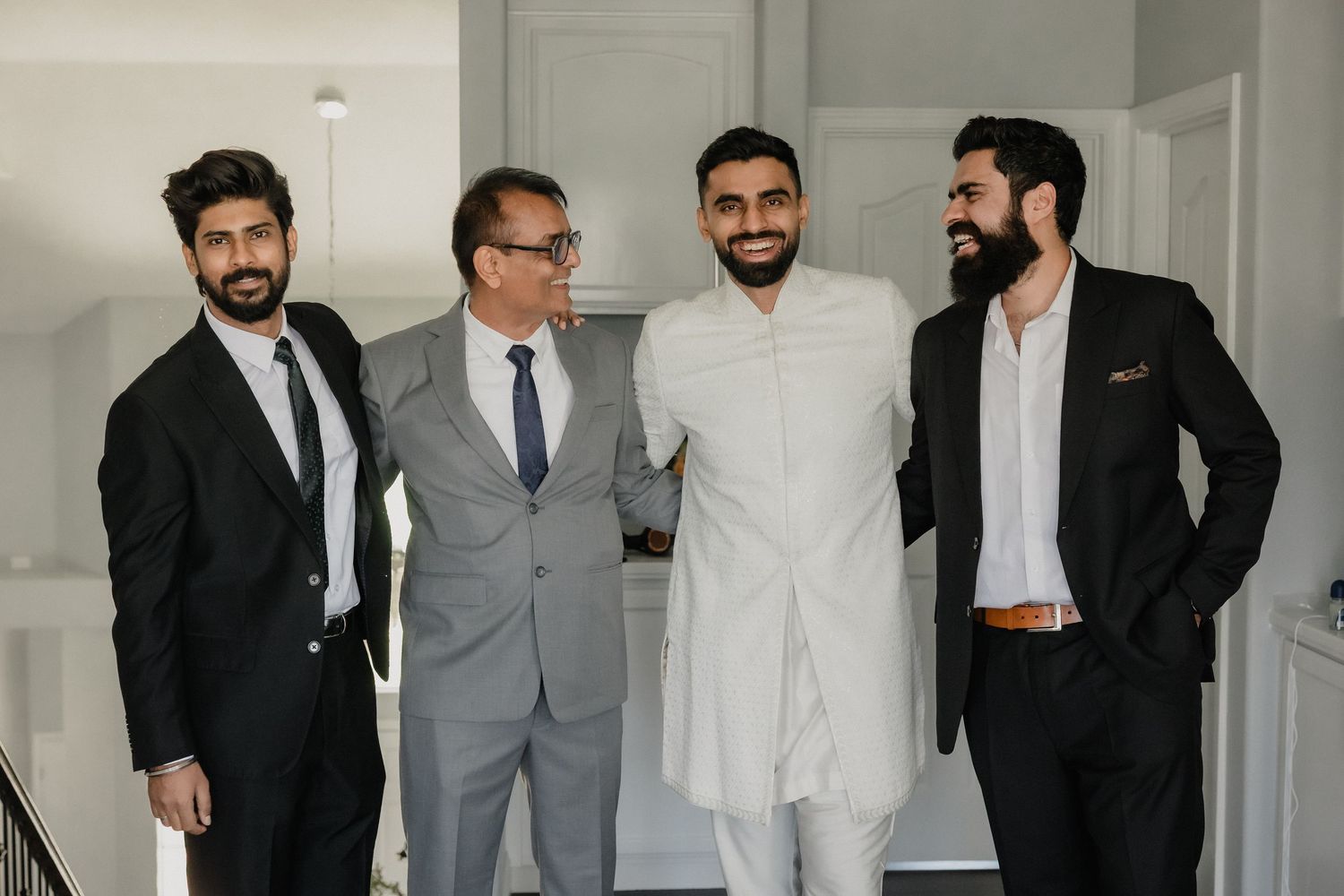 Groom in white sherwani poses with three groomsmen in dark suits on a staircase.
