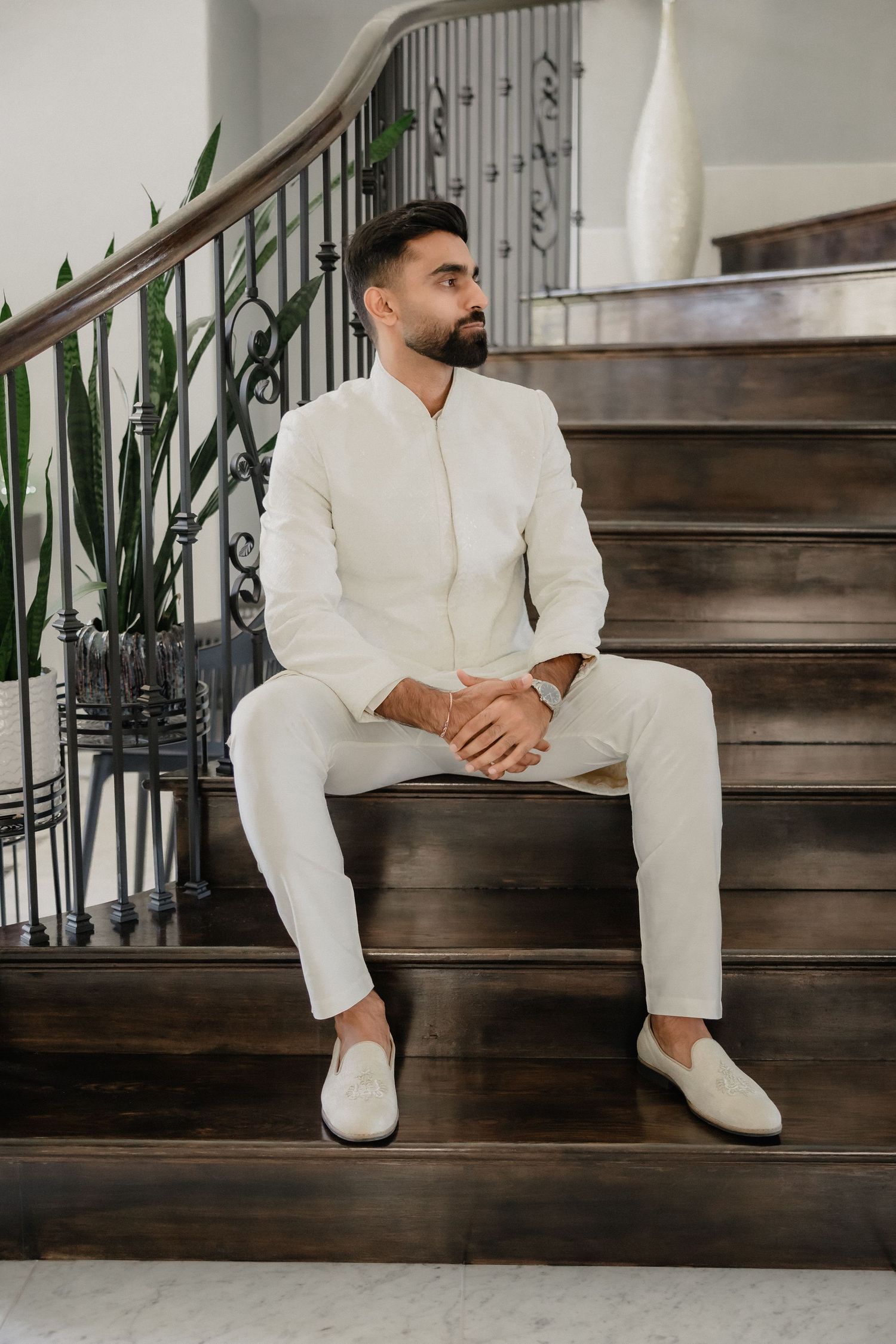 Bearded groom in white sherwani sits on wooden staircase in elegant venue before wedding ceremony.
