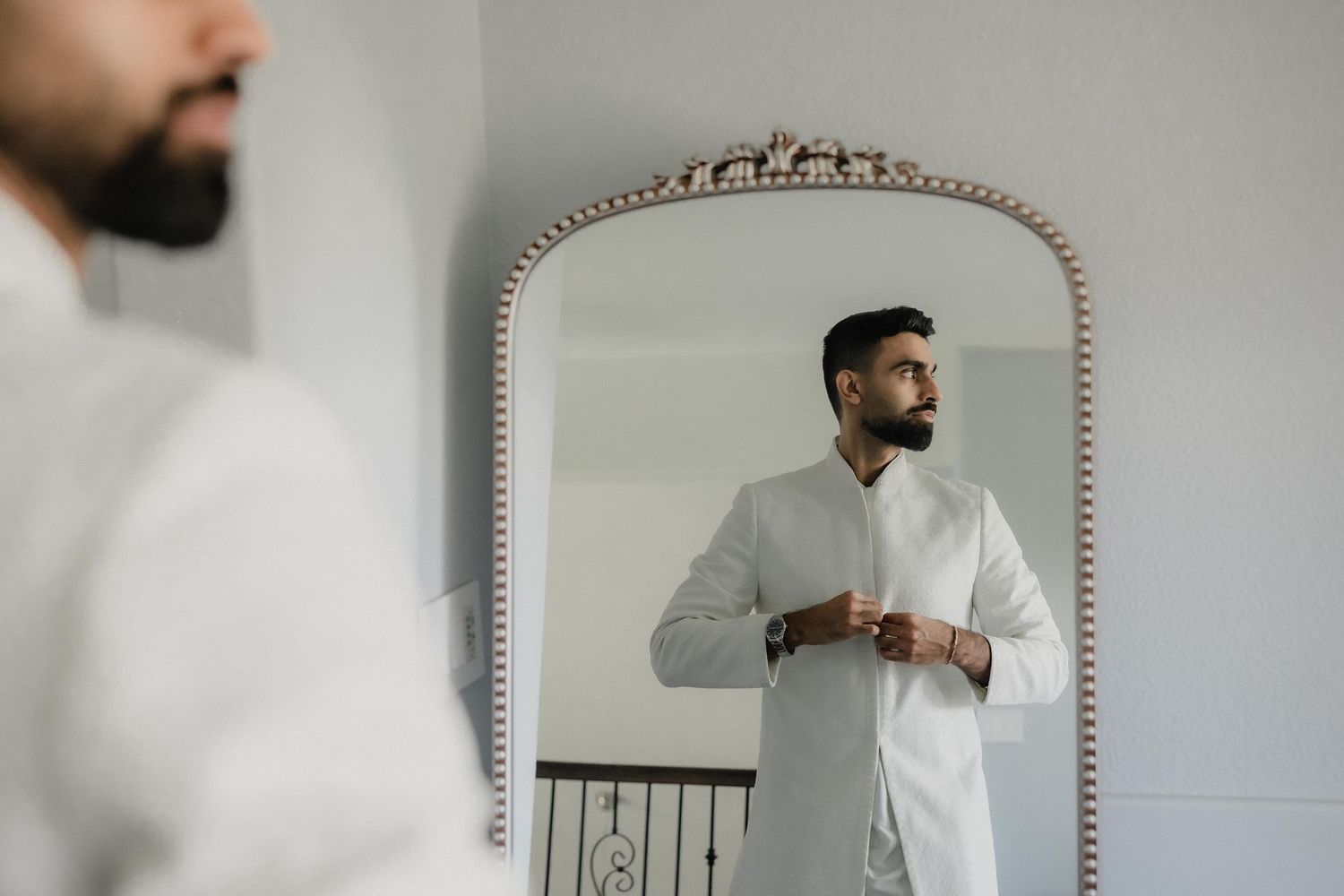 Groom in white sherwani adjusting his outfit while looking in an ornate mirror before his wedding ceremony.