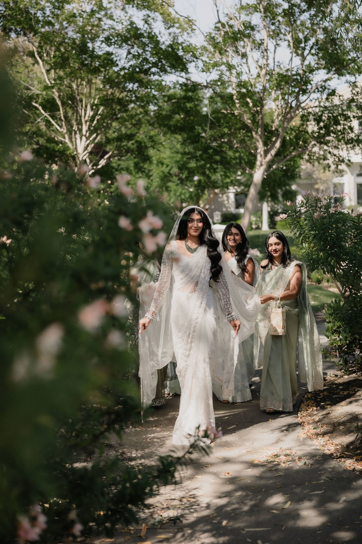 Bride in white saree walks with bridesmaids in sage green outfits along a garden path lined with lush trees.