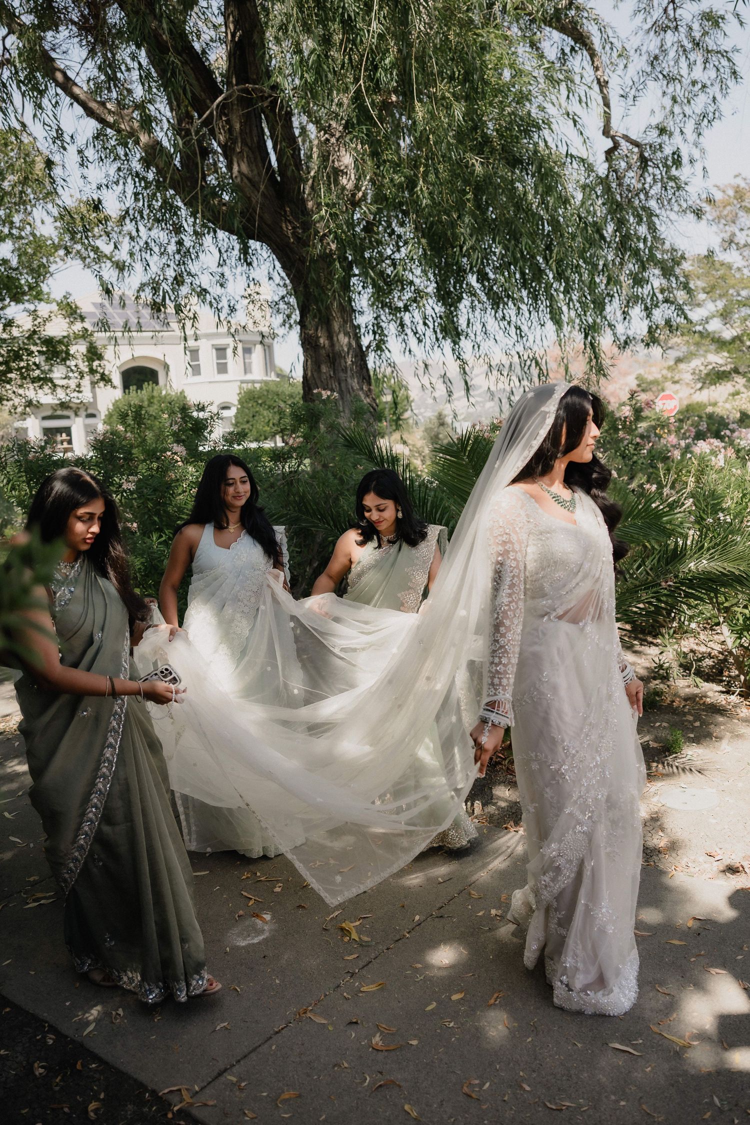 Bride in white lace saree and veil with bridesmaids in sage green sarees outdoors under trees.
