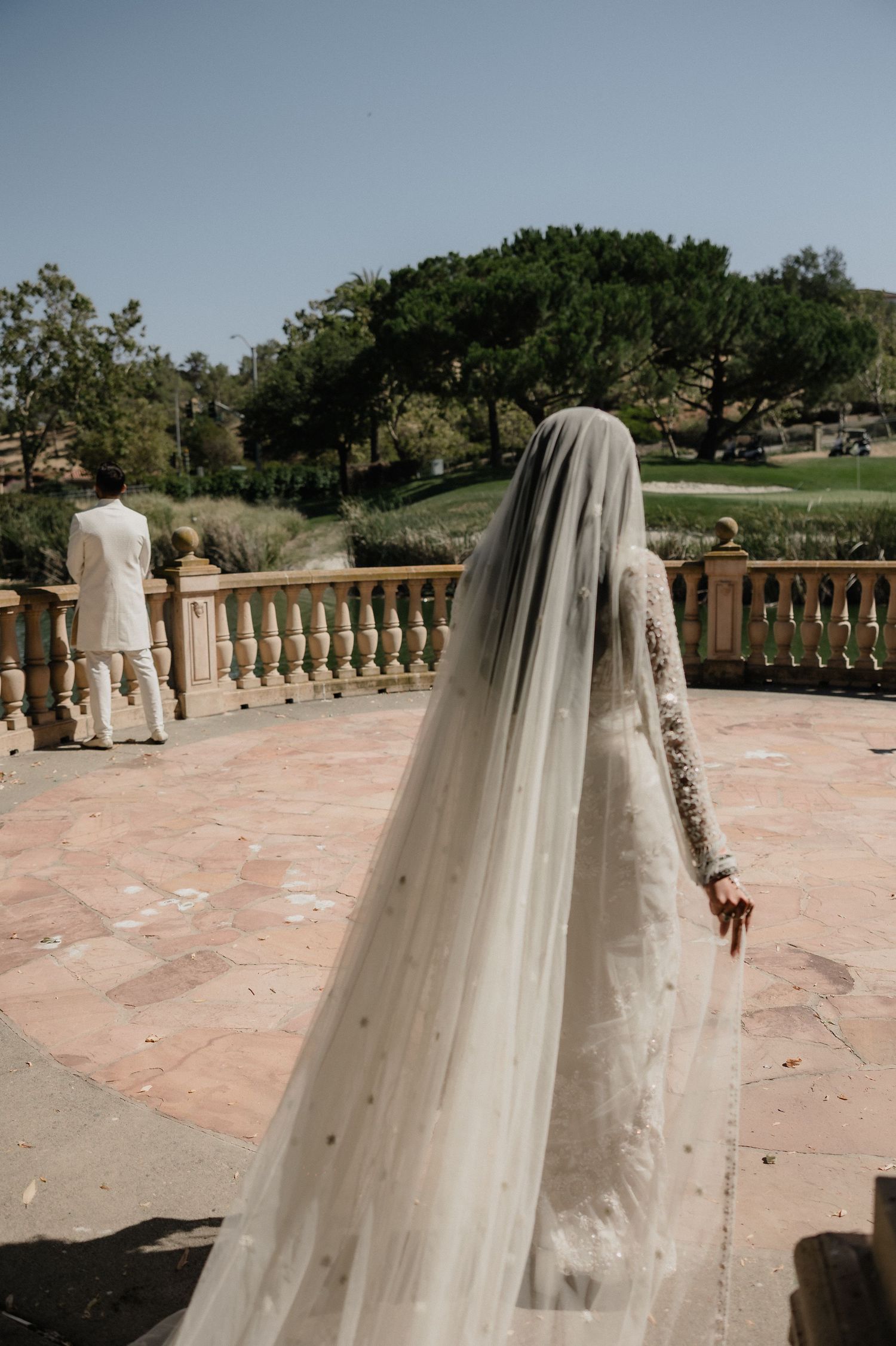 Bride in elegant white lace gown with long cathedral veil walking toward groom on a sunlit terrace.