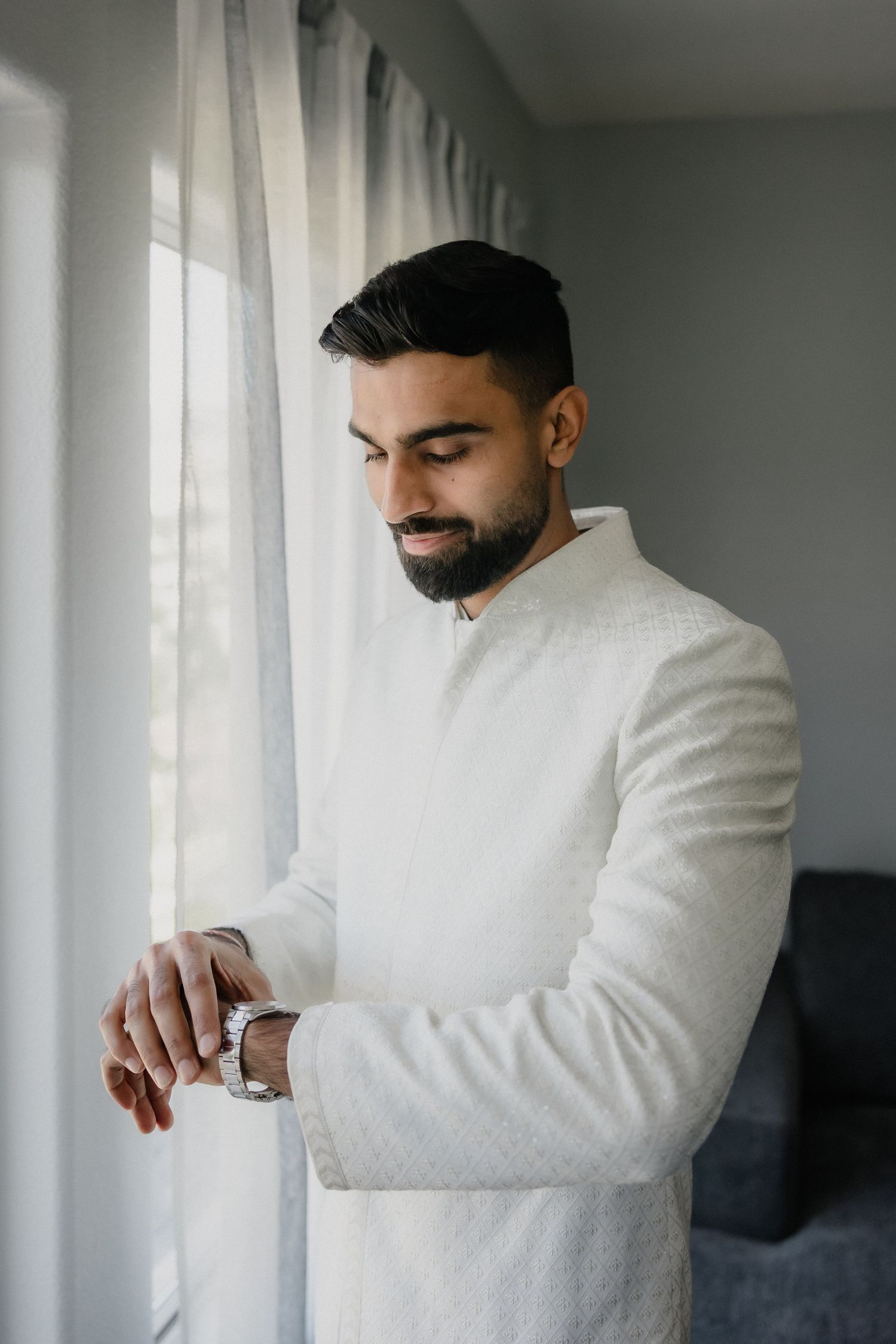 Groom in white sherwani checking his watch by a window on his wedding day.