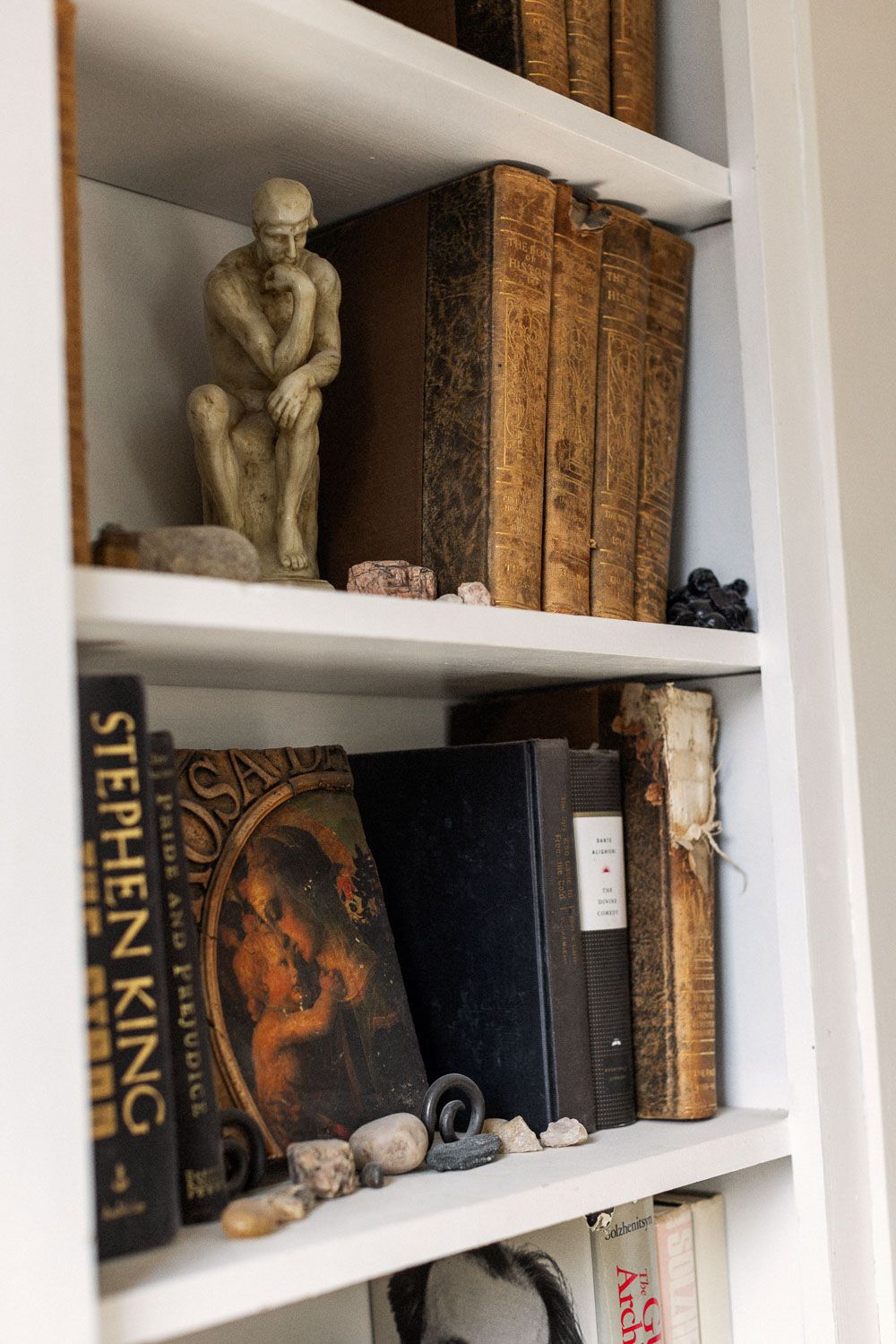 White bookshelf displaying a Rodin Thinker statue, antique books, and a Stephen King novel with decorative stones.