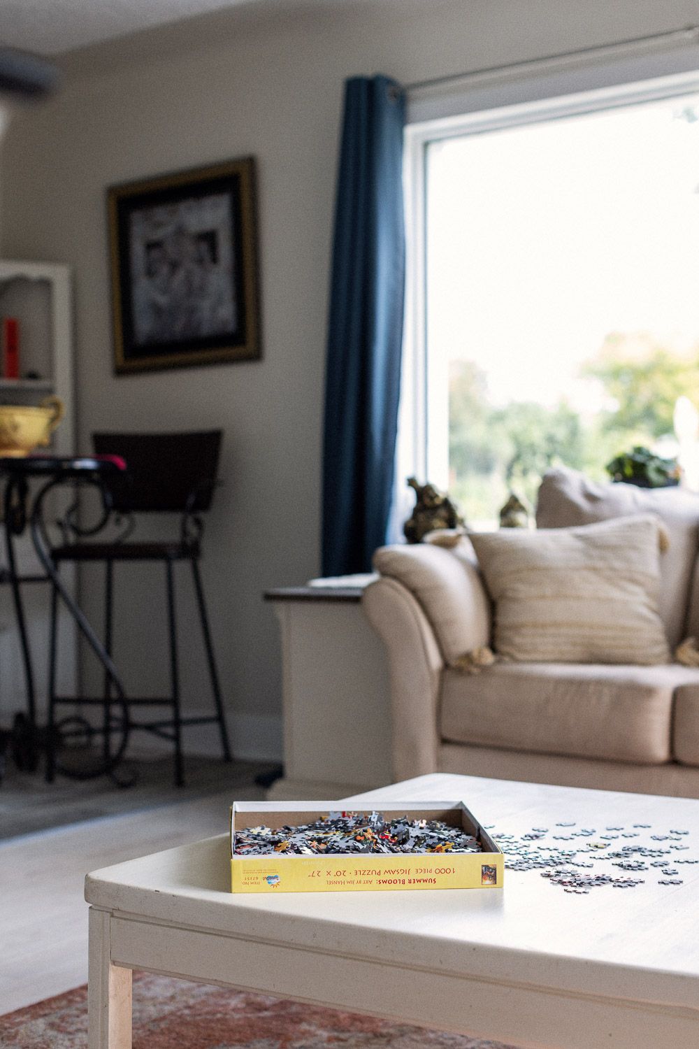 Cozy living room with a jigsaw puzzle on a white coffee table, beige sofa, blue curtains, and framed art on the wall.