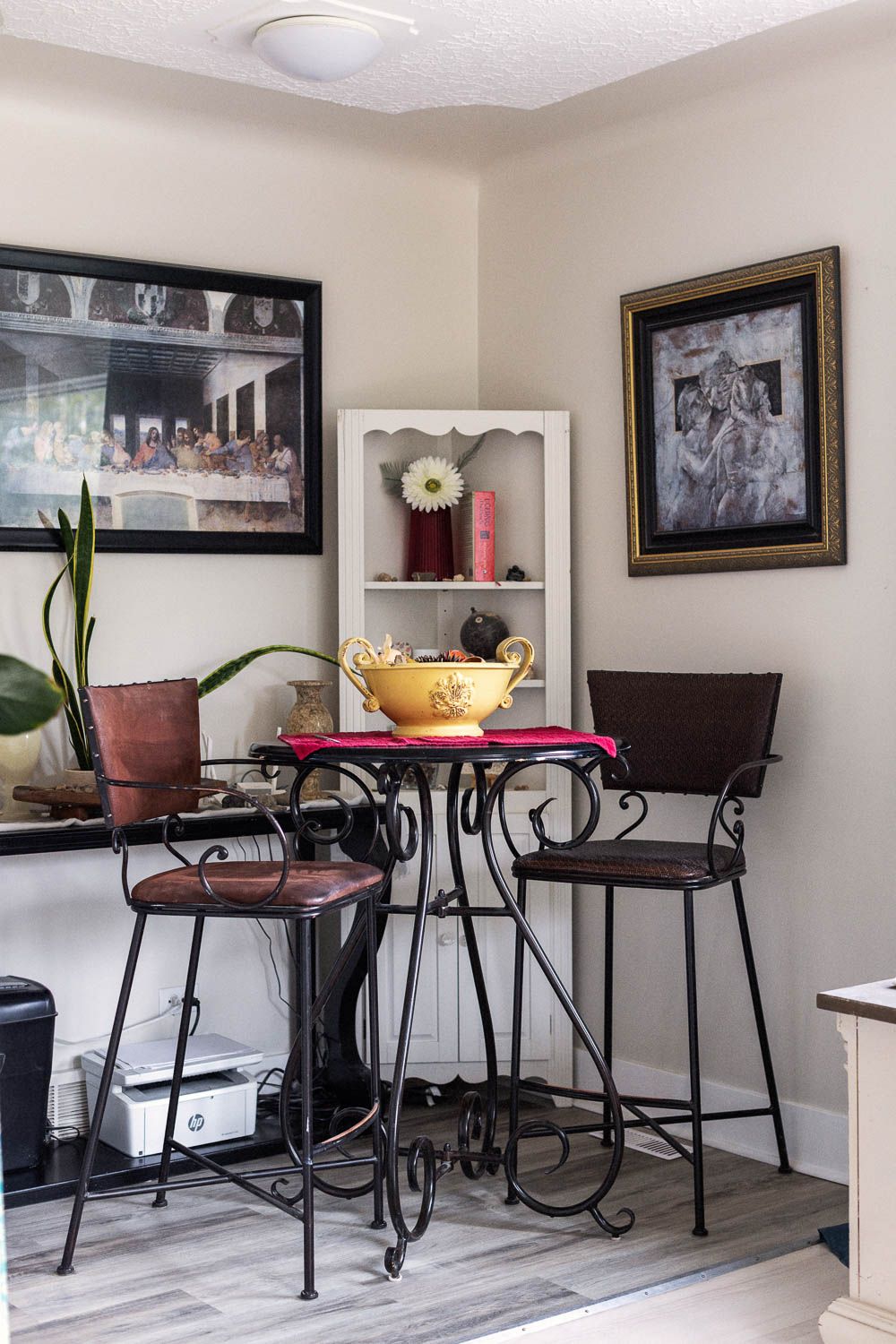 Cozy dining nook with wrought iron bistro set, yellow bowl centerpiece, and classic artwork including The Last Supper.