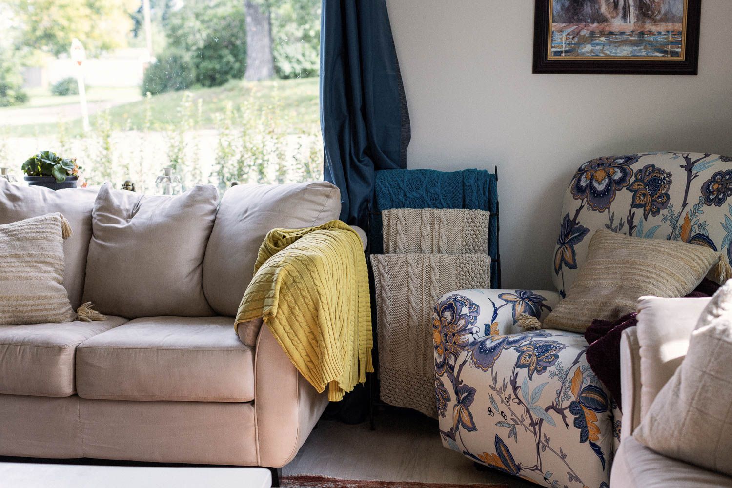 Cozy living room with beige sofa, yellow throw blanket, floral armchair, teal curtains, and cable knit pillows near a bright window.