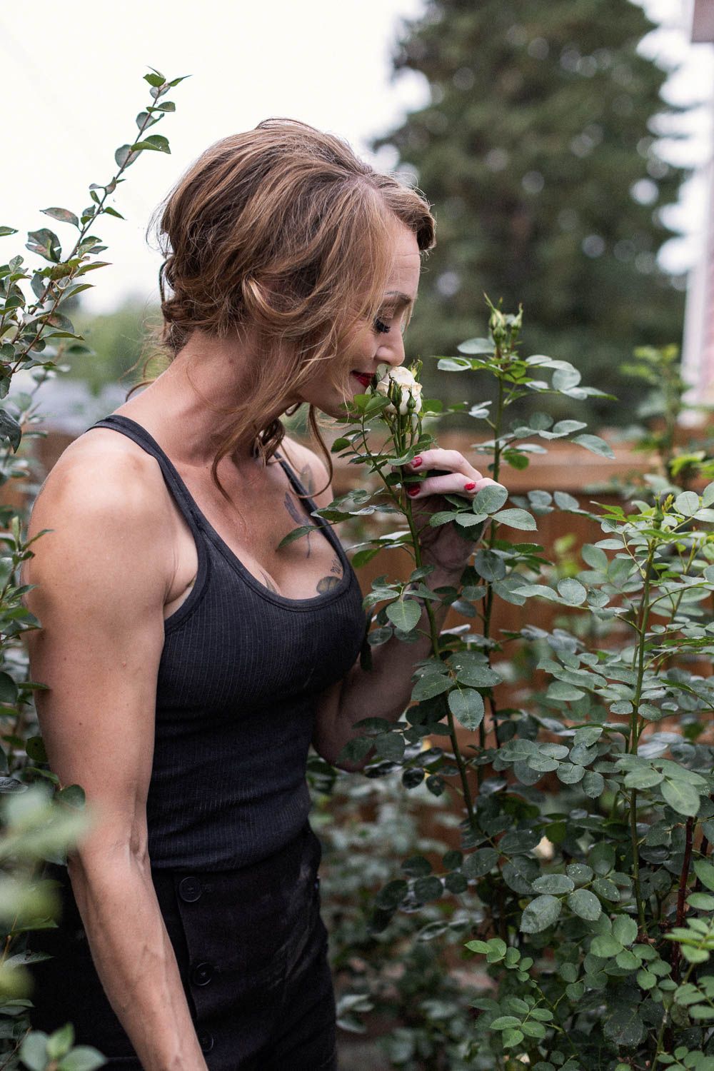 Woman in black tank top smelling a white rose in a lush garden setting.