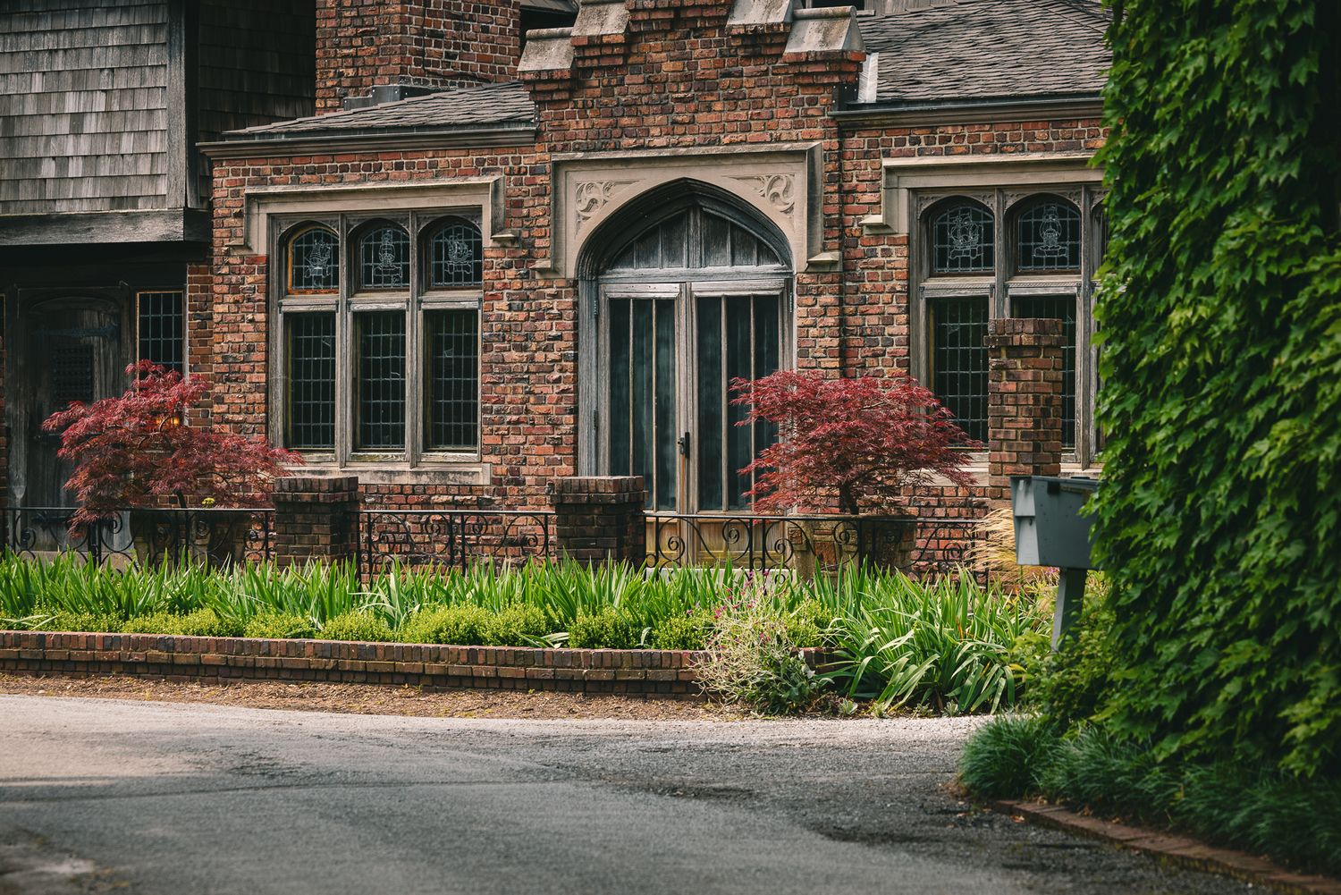Historic brick building with arched windows, lush greenery, red Japanese maples, and ivy-covered walls along a quiet street.