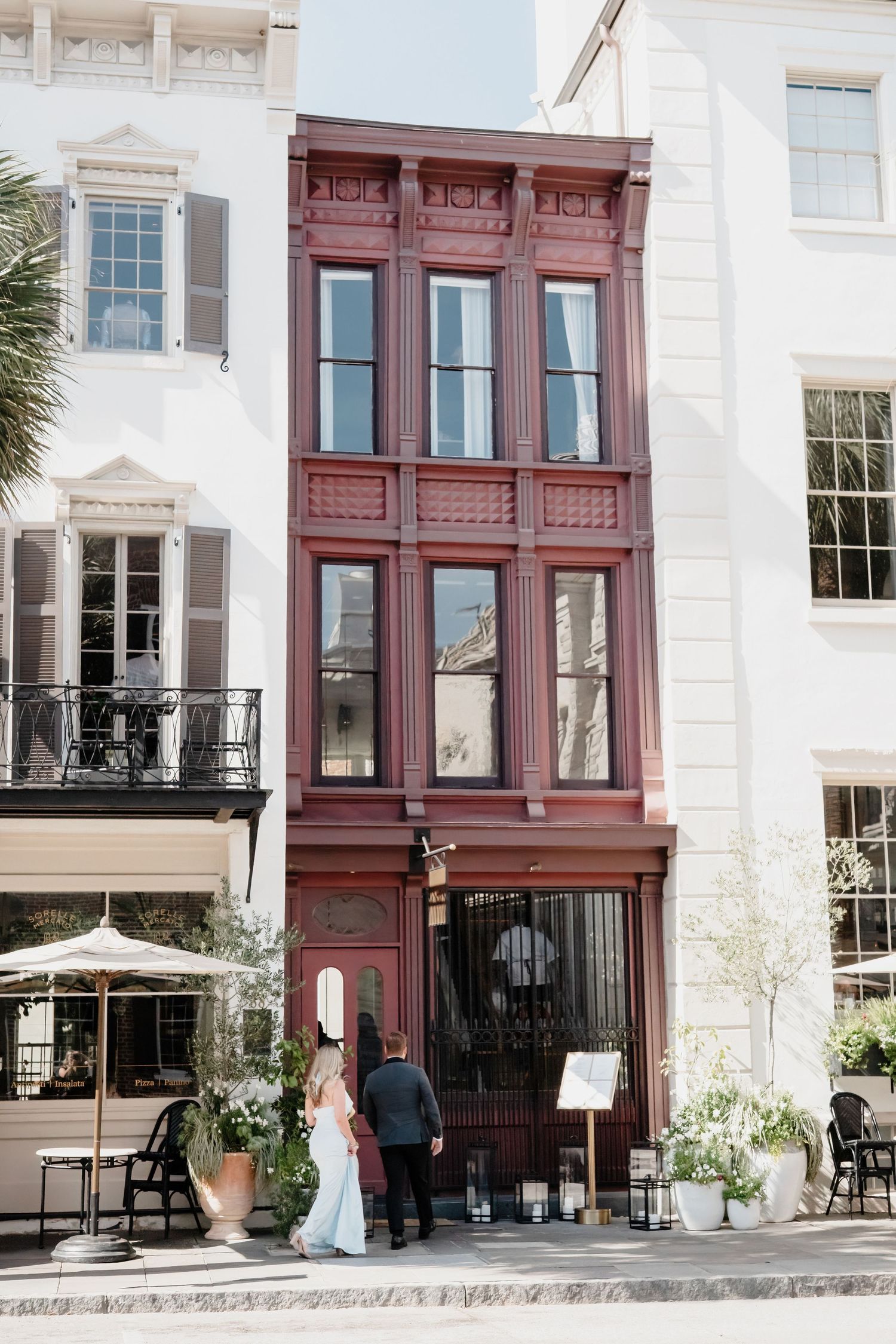 A bride and groom enter a historic red Victorian building facade on a sunny Charleston street.