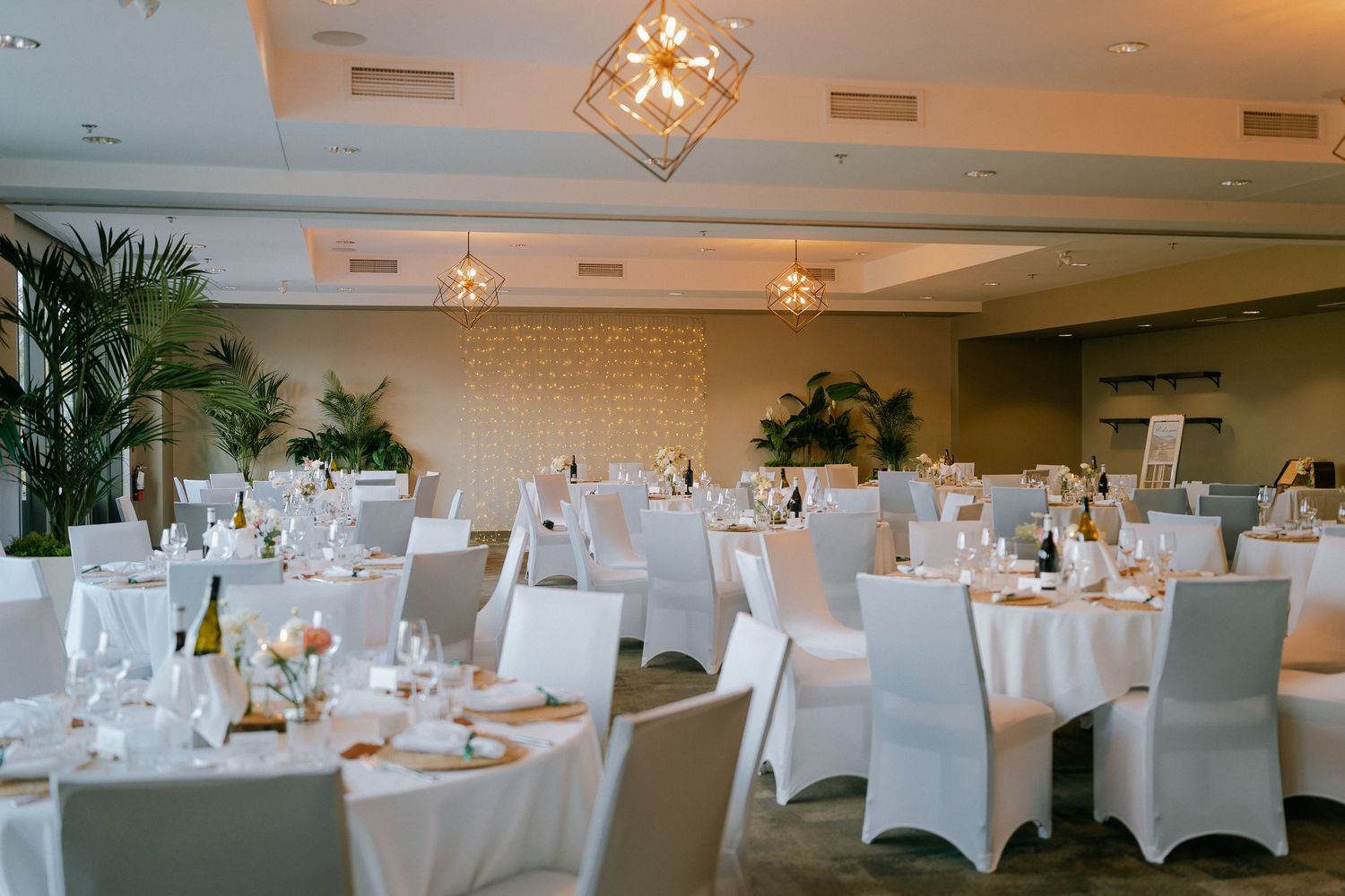 Elegant wedding reception hall with white-draped round tables, gold chargers, tropical plants, and crystal chandeliers.