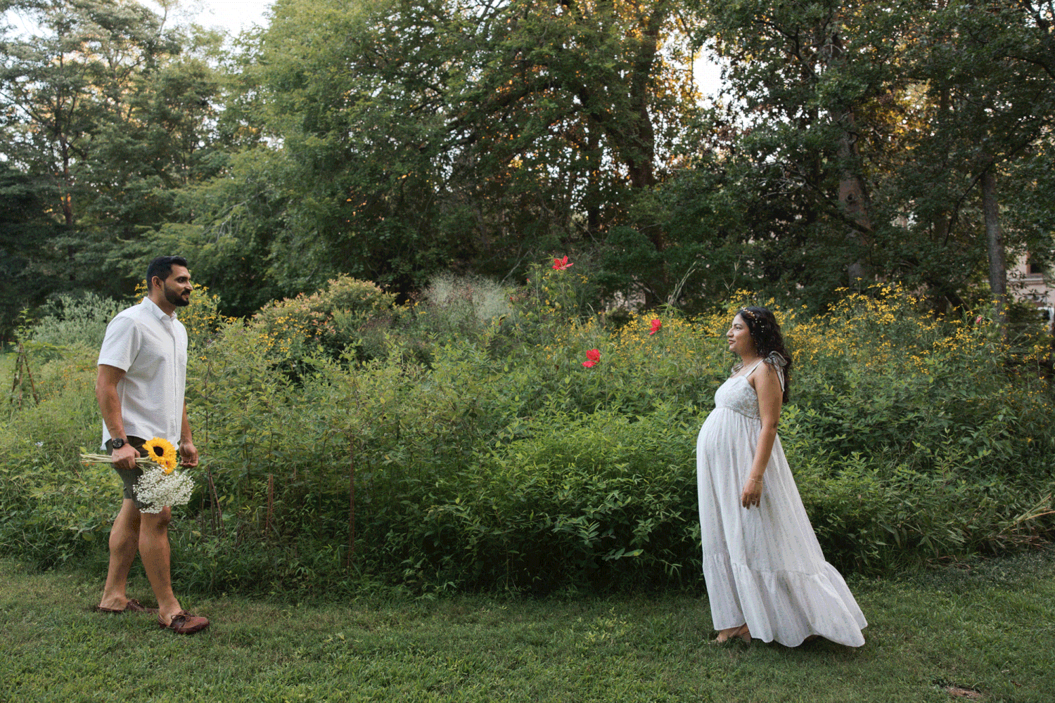 Pregnant woman in white dress and man holding sunflowers stand apart in a lush garden during maternity photoshoot.