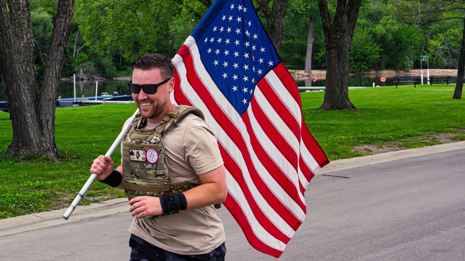 Smiling CrossFit athlete in a camo weight vest carries an American flag on the run leg of Murph, a tree-lined lake behind him.