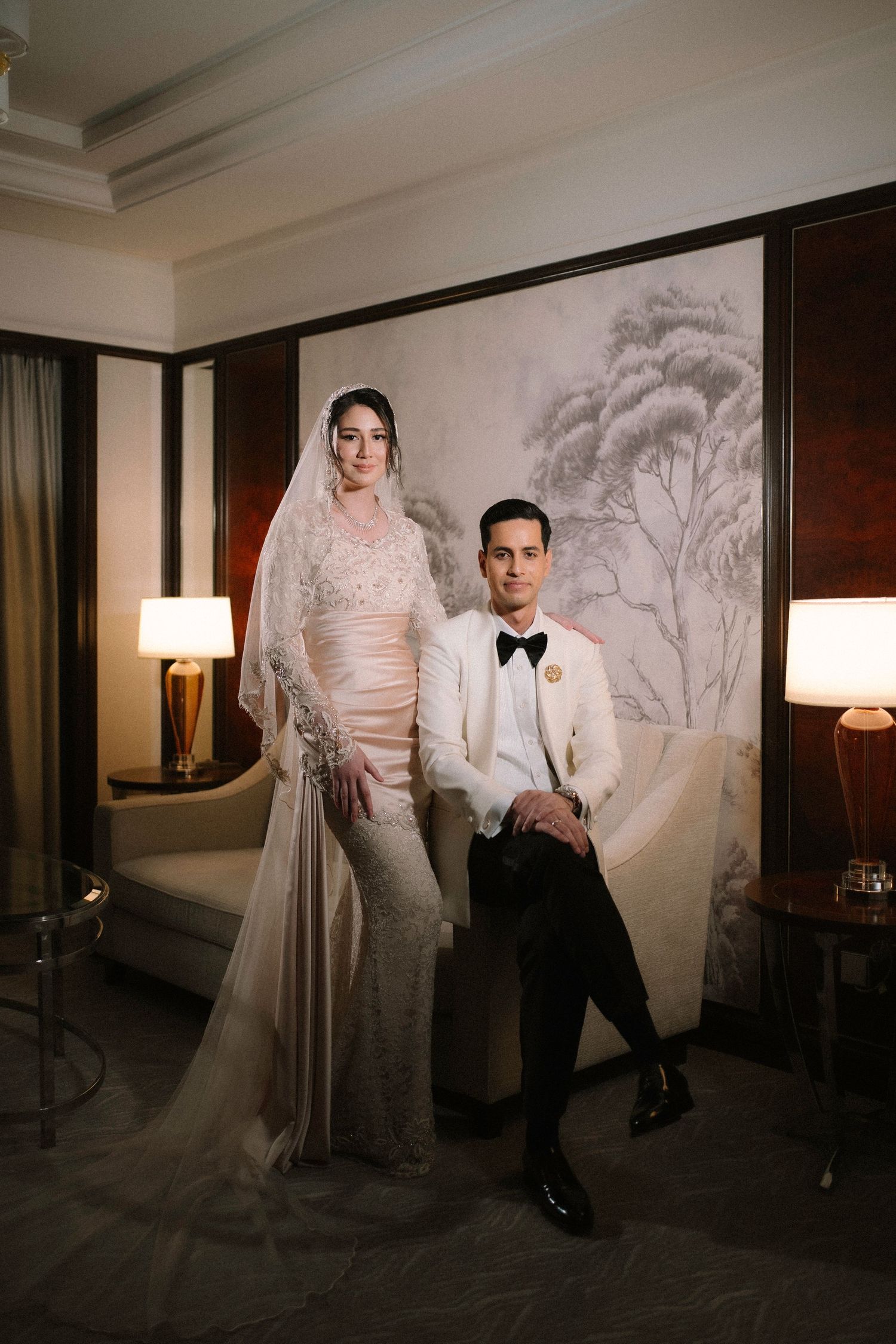Elegant bride in lace gown with veil stands beside groom in white tuxedo jacket in a luxurious hotel suite.