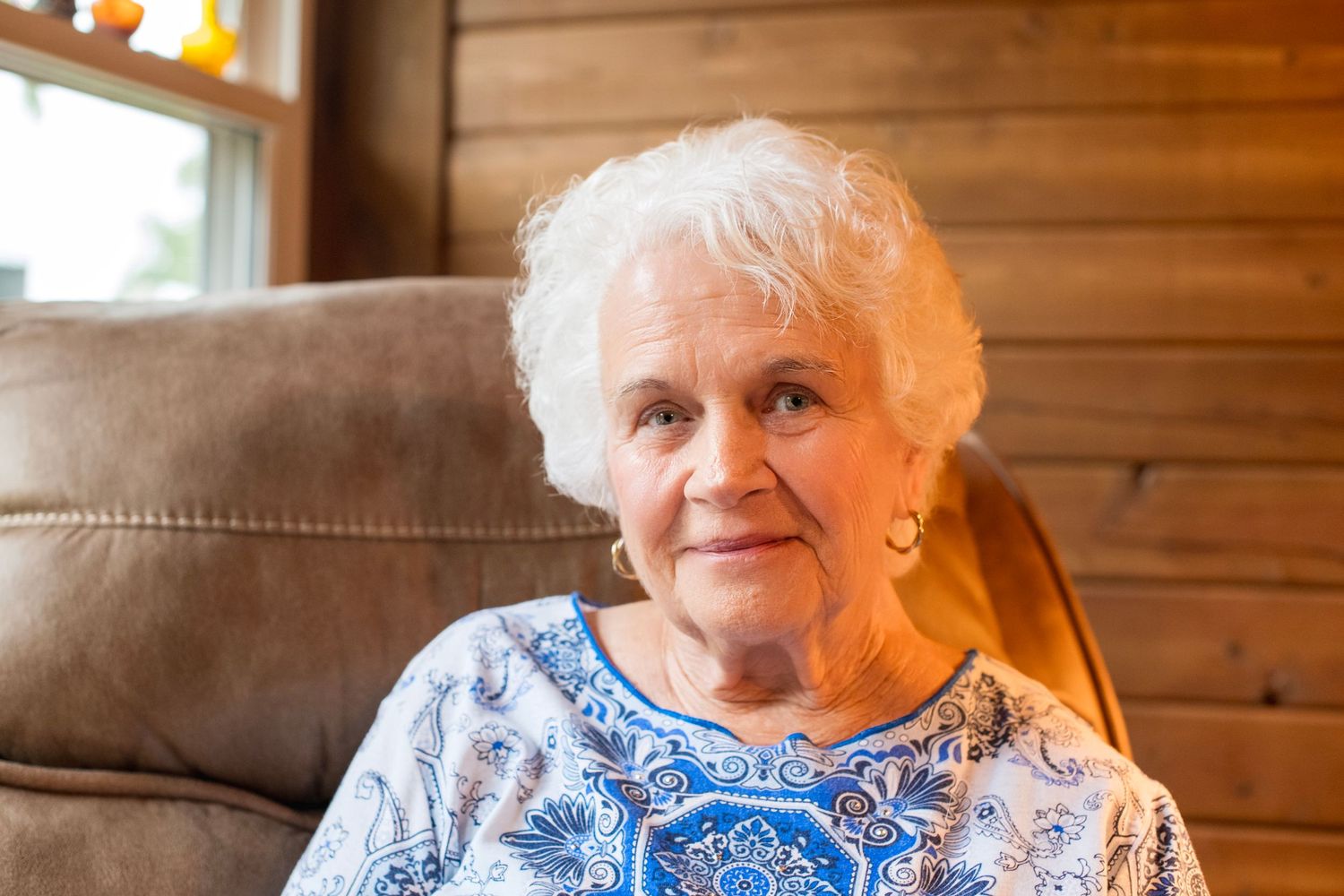 Elderly woman with white hair smiling warmly, seated in a brown leather chair wearing a blue floral blouse.