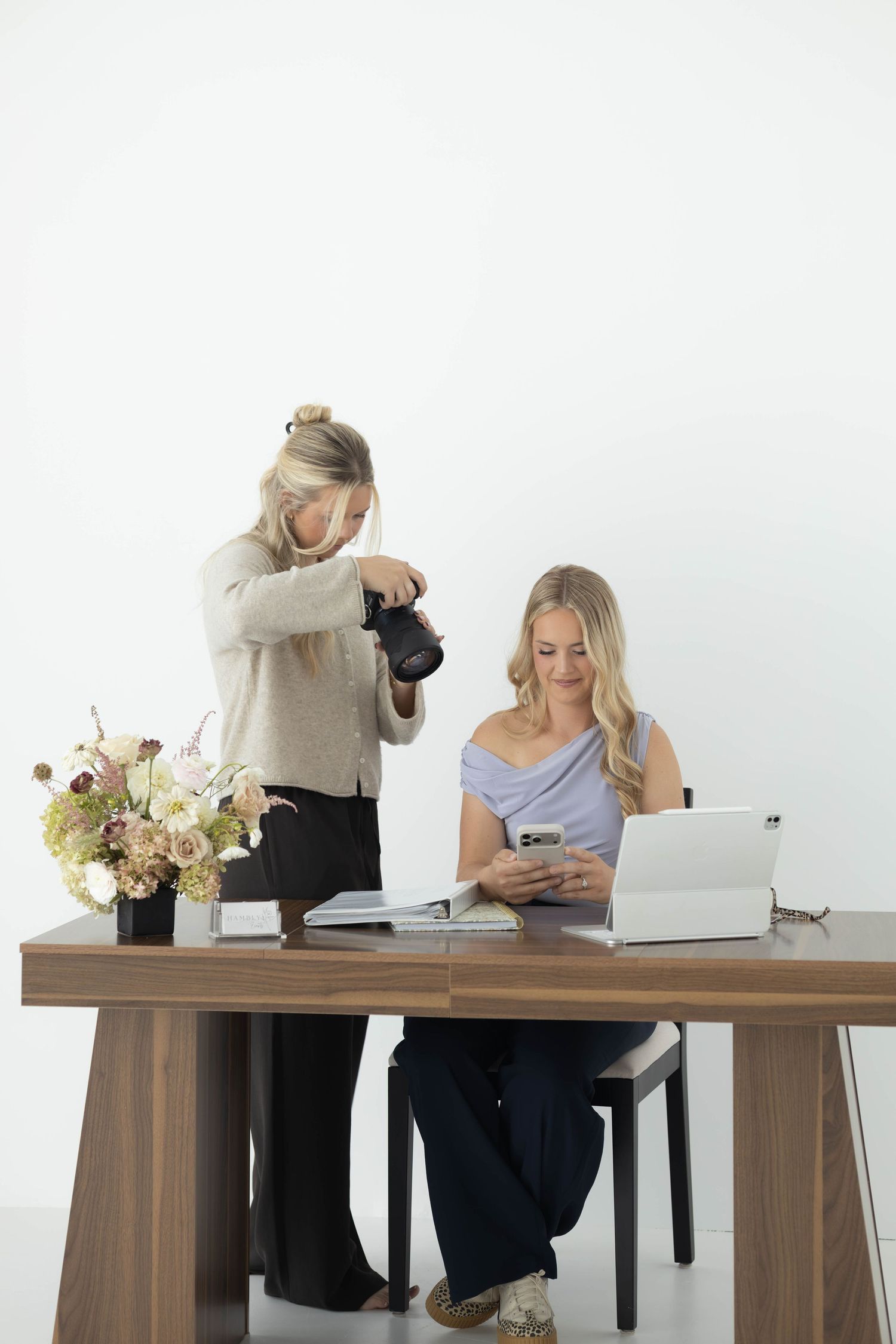 Two blonde women work at a desk with laptop, camera, and floral arrangement indoors.