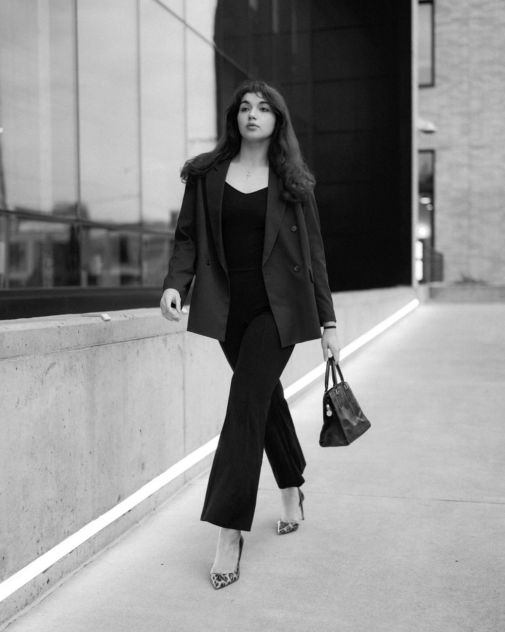 Woman in black blazer and wide-leg pants walks confidently outdoors, carrying a handbag, wearing leopard heels.