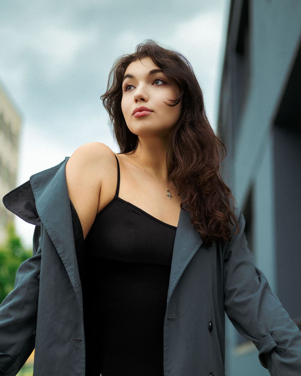 Young woman in black dress and gray jacket poses outdoors with cloudy sky backdrop.