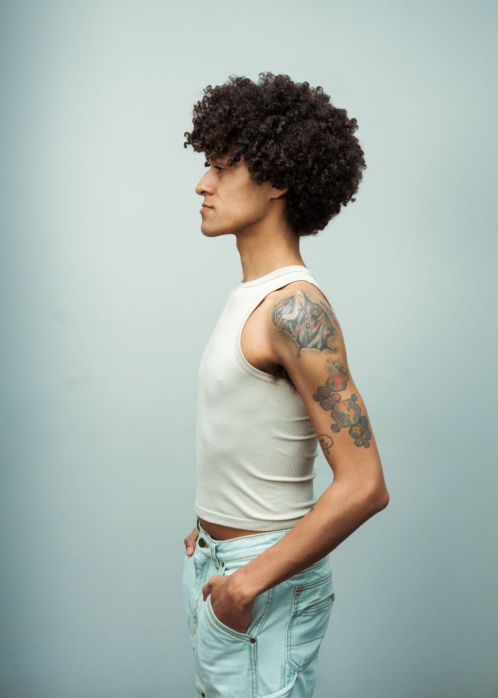 Person with curly hair and arm tattoos wearing a white crop top and light jeans, posed sideways against a blue background.