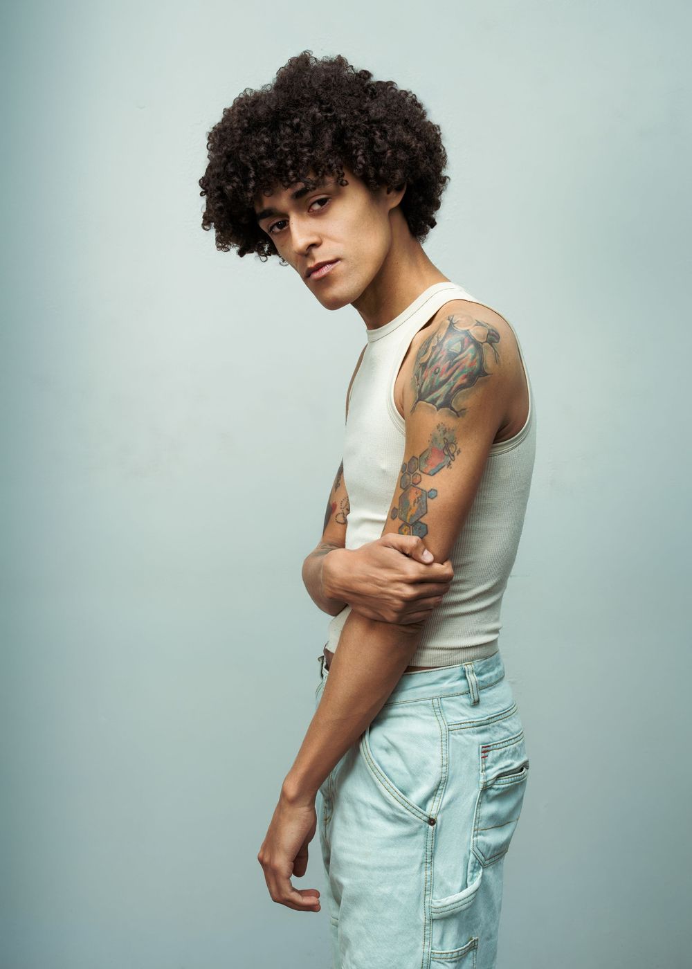 Young man with curly afro hair, tattoos on arm, wearing white sleeveless top and light blue jeans, posing sideways.