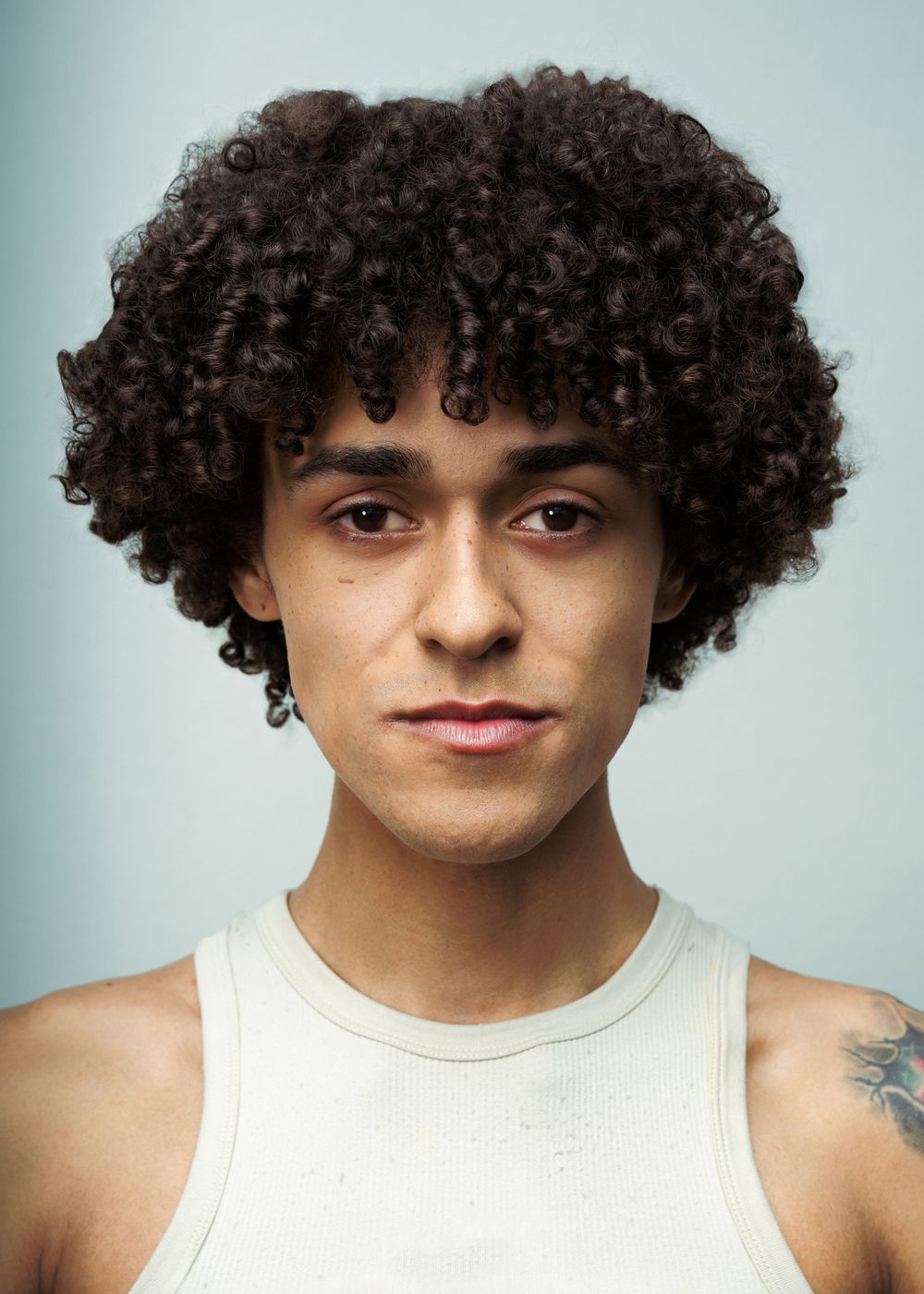 Young man with dark curly afro hair wearing a white tank top with a tattoo on his shoulder, against a light blue background.