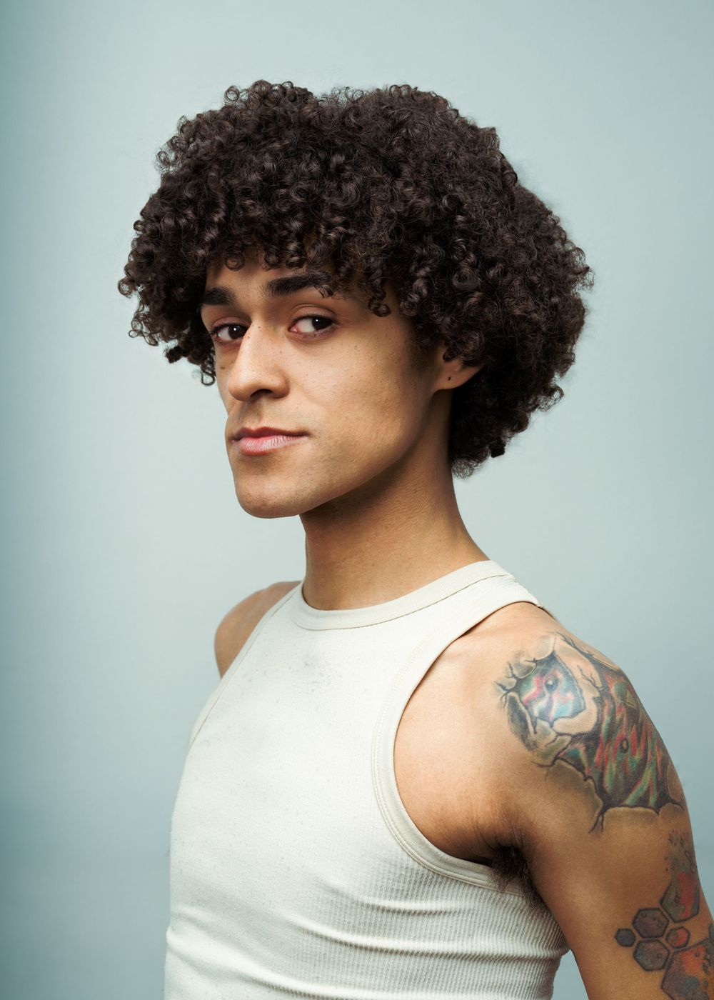 Young man with curly afro hair wearing a white tank top, showcasing a colorful tattoo on his shoulder.