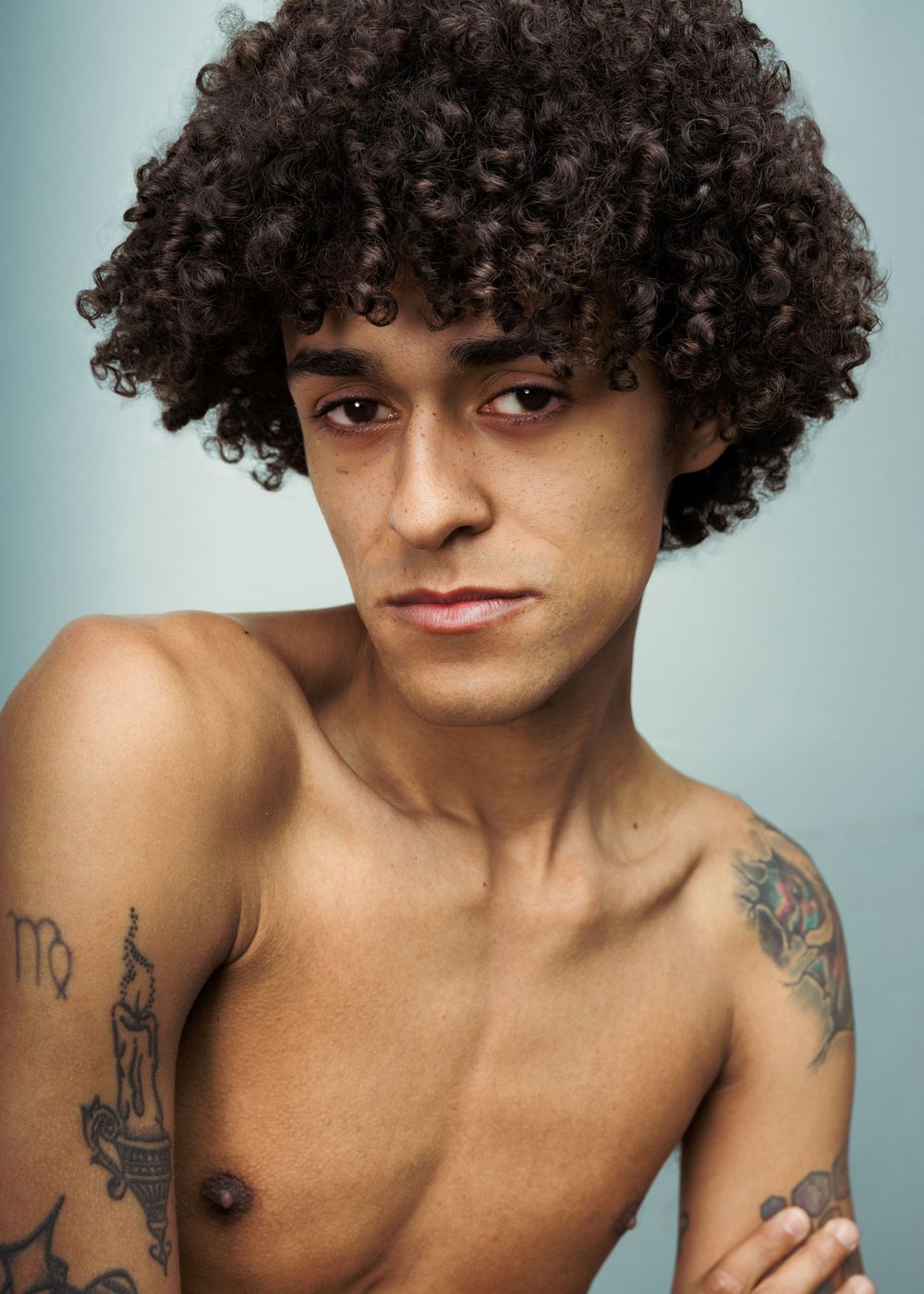 Young shirtless man with curly afro hair displaying multiple tattoos on his arms and chest.