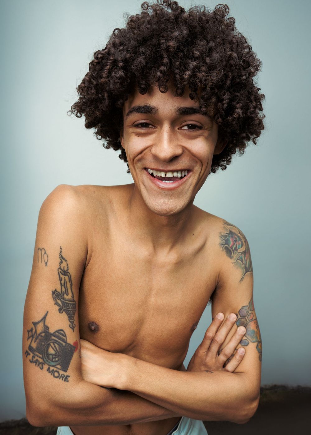 Smiling young man with curly afro hair, shirtless with tattoos on both arms, arms crossed, light blue background.