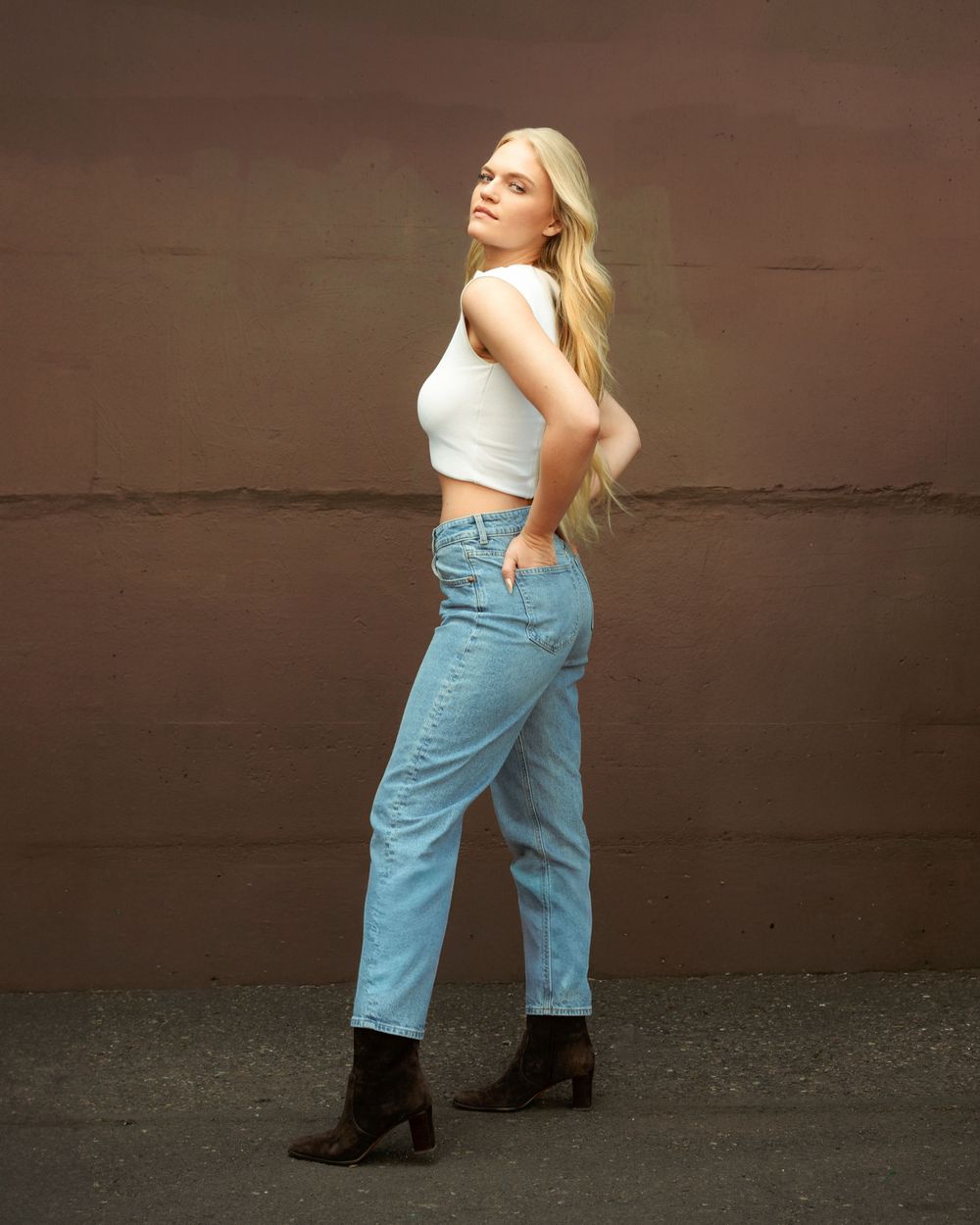 Blonde woman posing in white crop top, light blue jeans, and brown ankle boots against a brown wall.